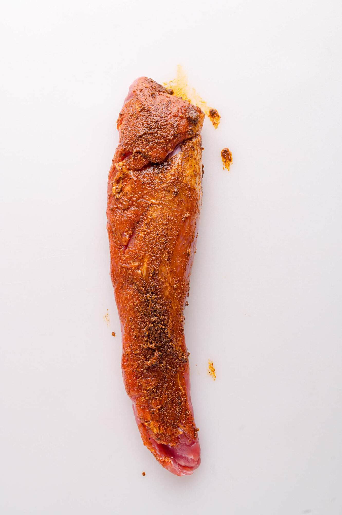 A raw pork tenderloin coated with a reddish-brown spice rub sits on a white surface.