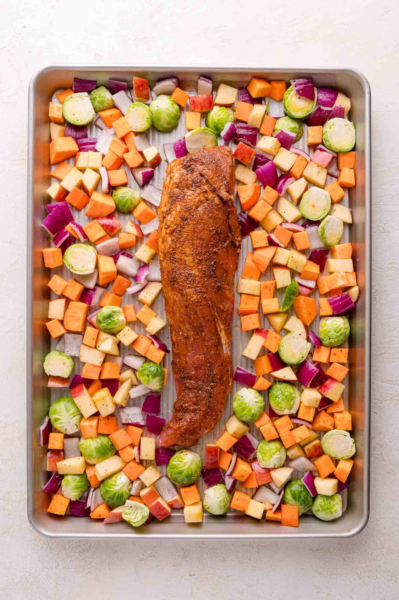 A sheet pan with a seasoned pork tenderloin in the center, surrounded by diced sweet potatoes, Brussels sprouts, red onions, and apples.