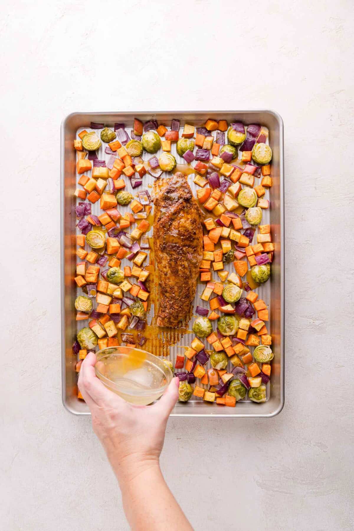 A hand holds a bowl of maple syrup and mustard mixture above a baking sheet with roasted pork tenderloin, sweet potatoes, Brussels sprouts, and red onions.
