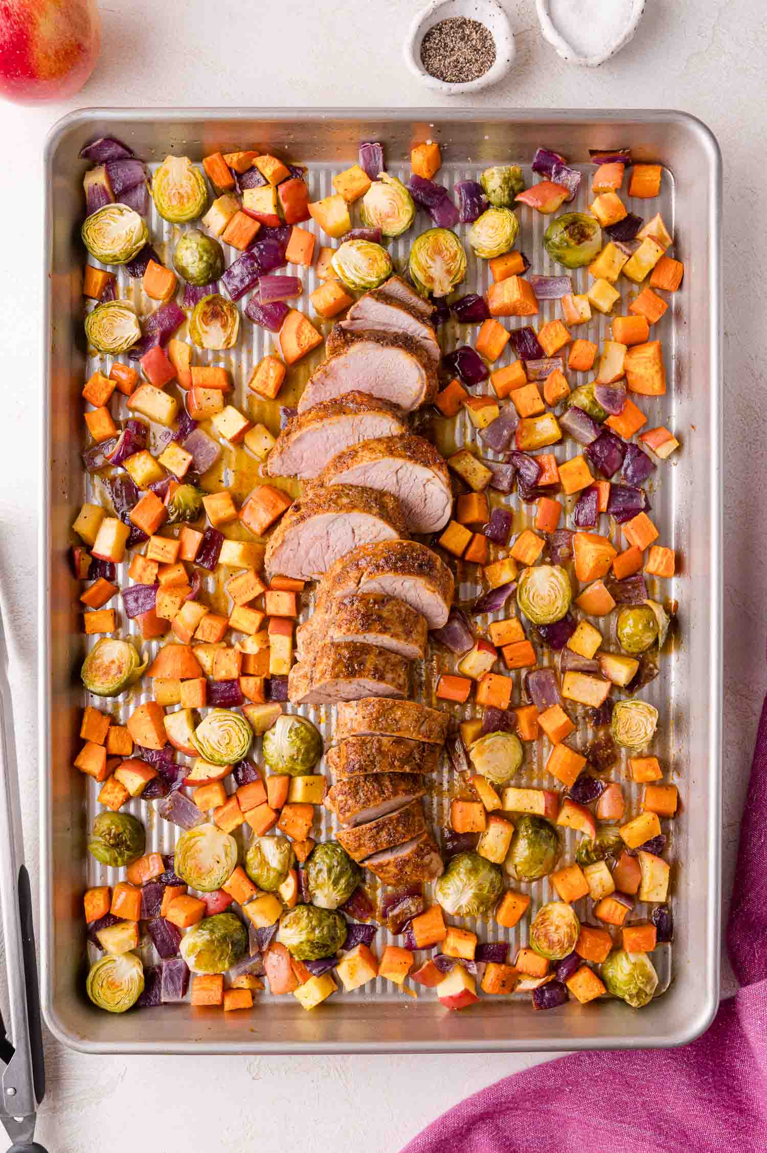 A baking sheet with sliced pork tenderloin surrounded by roasted Brussels sprouts, sweet potatoes, apples, and red onions. Salt and pepper bowls are nearby.
