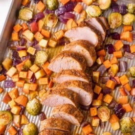 Sliced pork tenderloin with roasted Brussels sprouts, red onions, sweet potatoes, and root vegetables on a baking sheet.