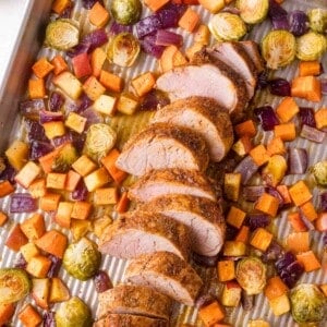 Sliced pork tenderloin with roasted Brussels sprouts, red onions, sweet potatoes, and root vegetables on a baking sheet.