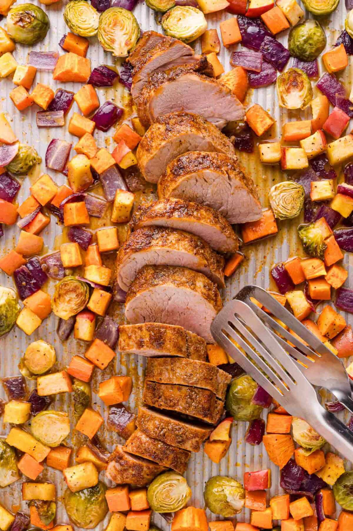 Sliced pork tenderloin on a baking sheet surrounded by roasted sweet potatoes, Brussels sprouts, and red onions, with metal tongs beside the pork.