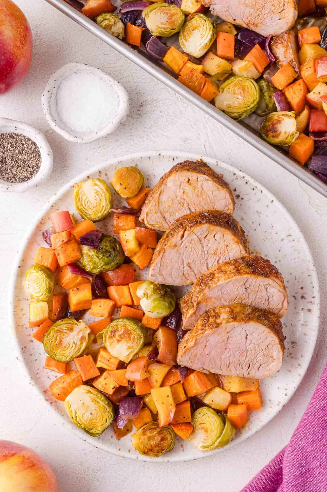 A plate with three slices of cooked pork tenderloin served alongside roasted Brussels sprouts, sweet potatoes, onions, and apples. A baking sheet with more food is in the background.