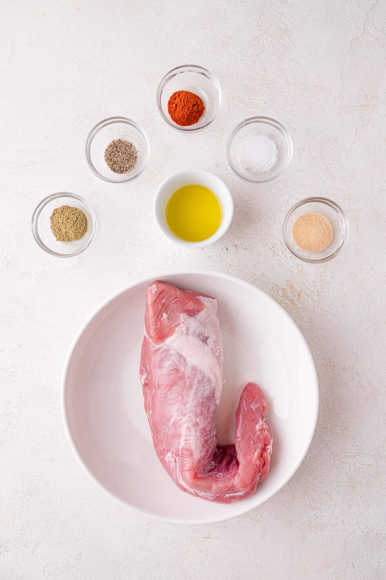Raw pork tenderloin in a white bowl surrounded by bowls of olive oil, salt, pepper, paprika, garlic powder, and dried herbs on a light surface.