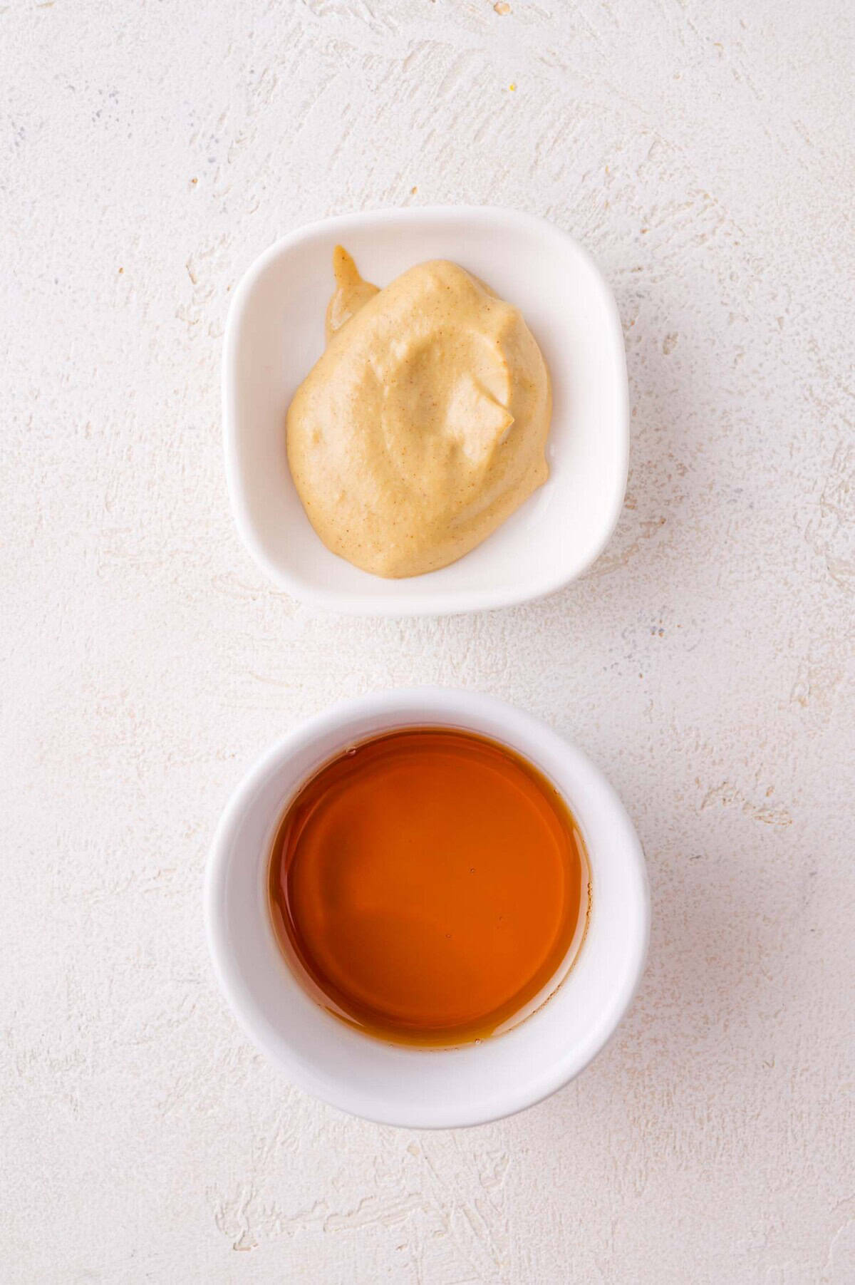 A small white bowl of creamy dijon mustard above a small white bowl of maple syrup, both on a light textured surface.