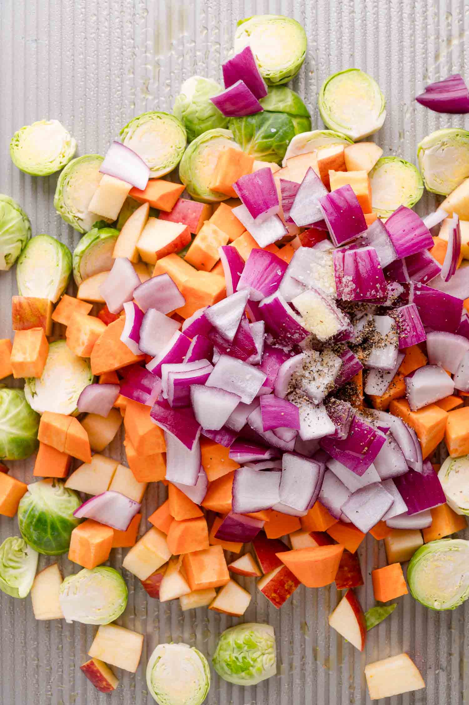 Chopped Brussels sprouts, red onion, sweet potato, and apples, on a baking sheet, sprinkled with salt and pepper.