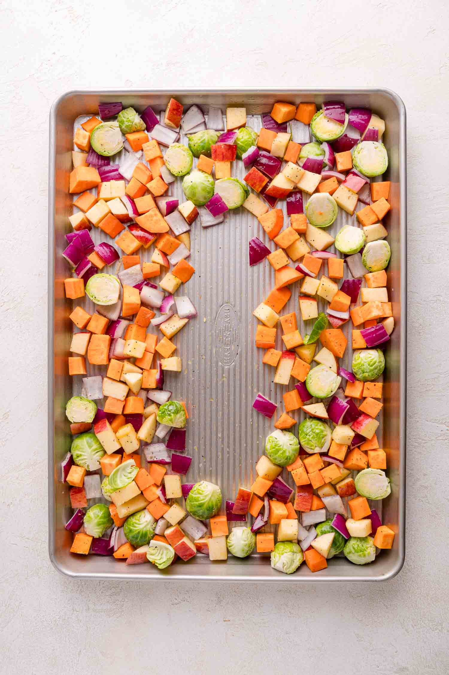 Chopped sweet potatoes, Brussels sprouts, red onions, and aples spread on a metal baking sheet, ready for roasting.