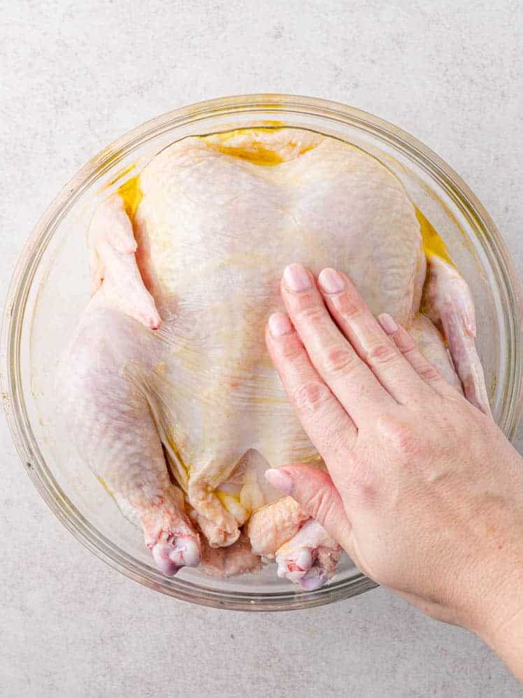 A hand rubbing oil onto a whole raw chicken in a glass bowl on a light-colored surface.