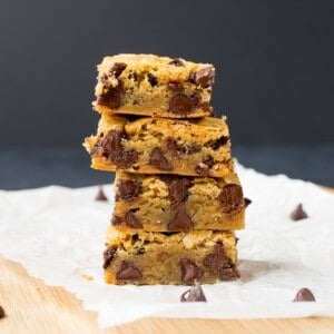 A stack of four chocolate chip blondies sits on a sheet of parchment paper, with a few scattered chocolate chips around them.