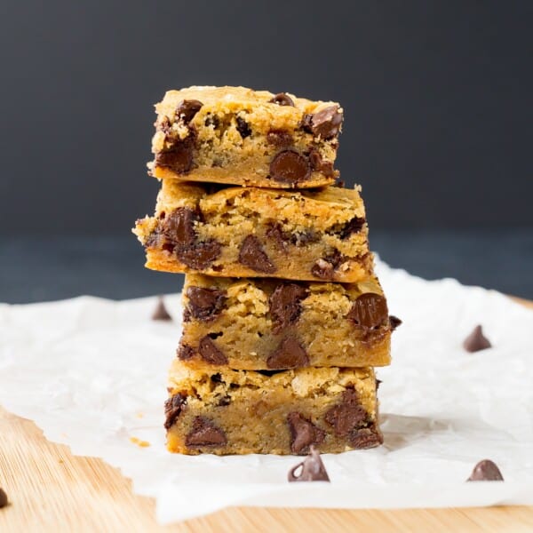 A stack of four chocolate chip blondies sits on a sheet of parchment paper, with a few scattered chocolate chips around them.