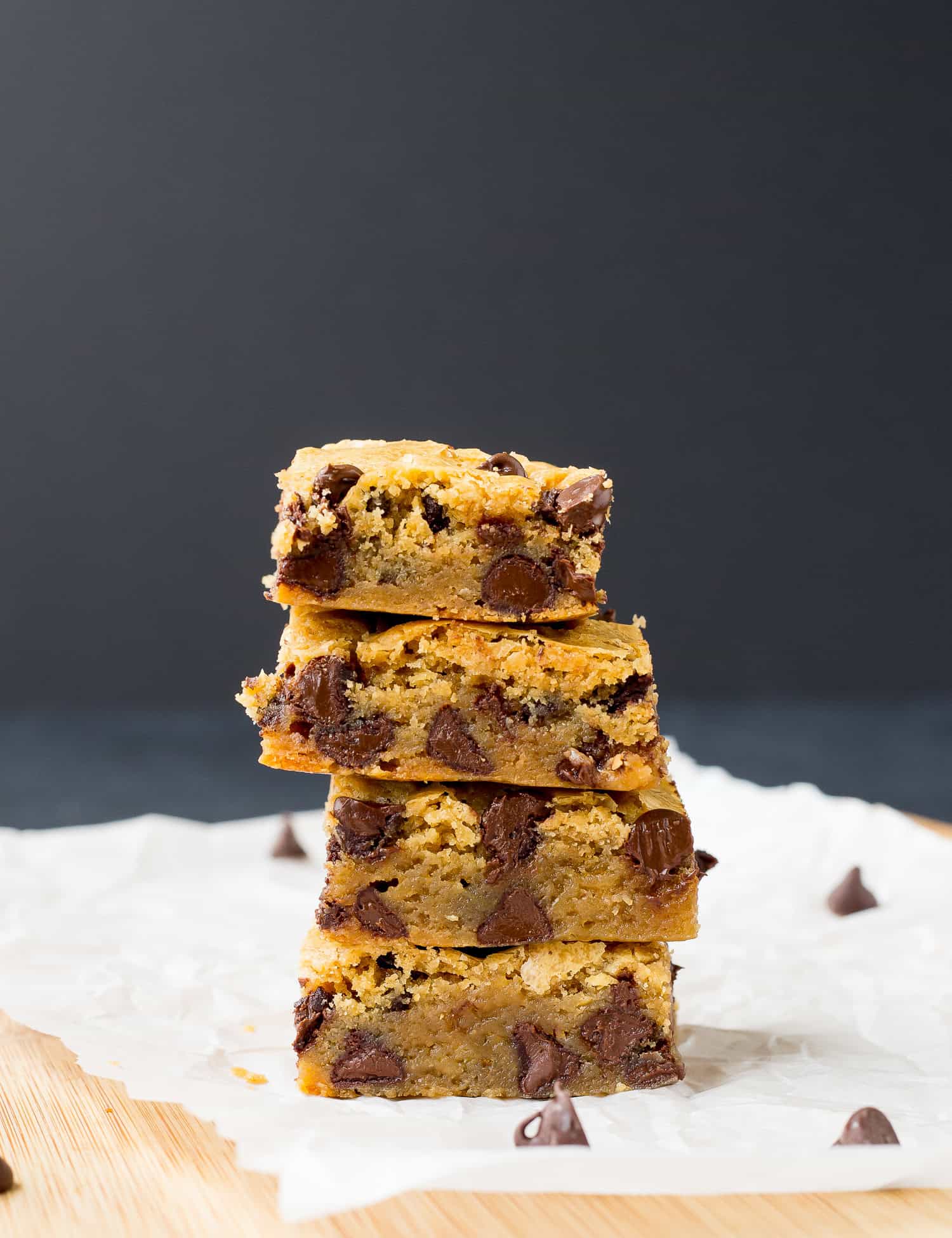 A stack of four chocolate chip blondies on parchment paper with scattered chocolate chips on a wooden surface.