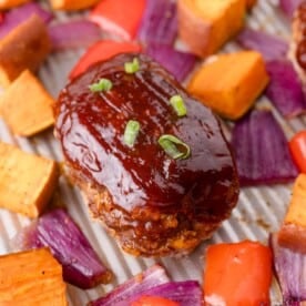 Mini meatloaf topped with barbecue sauce and green onion, surrounded by roasted sweet potatoes, red onions, and red bell peppers on a sheet pan.