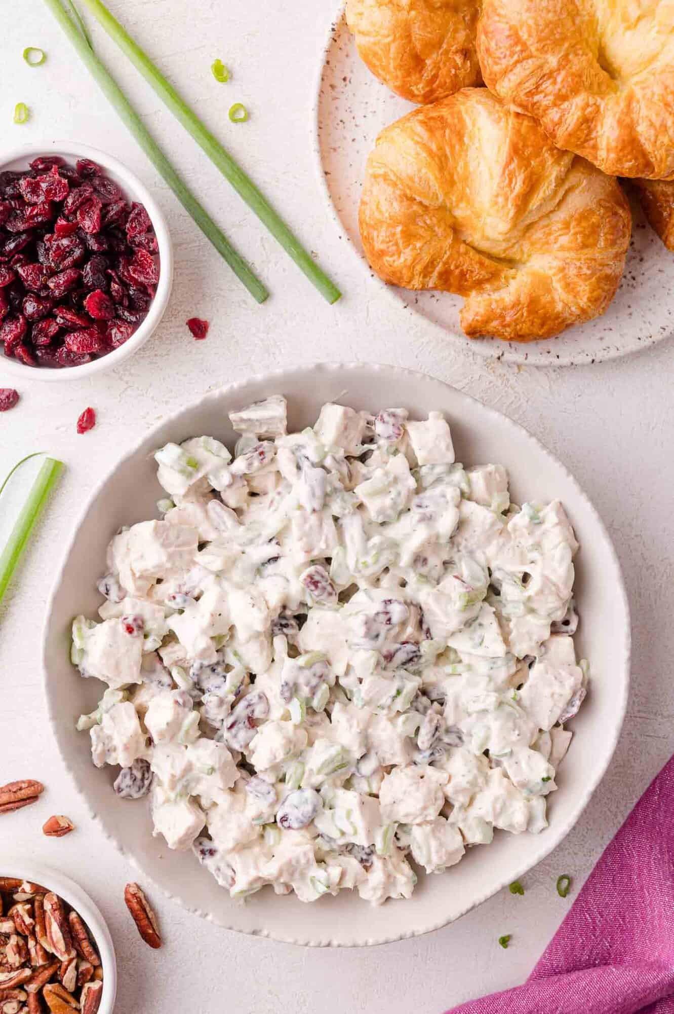 A bowl of chicken salad with celery and dried cranberries sits on a white table next to croissants, a bowl of dried cranberries, pecans, and green onions.