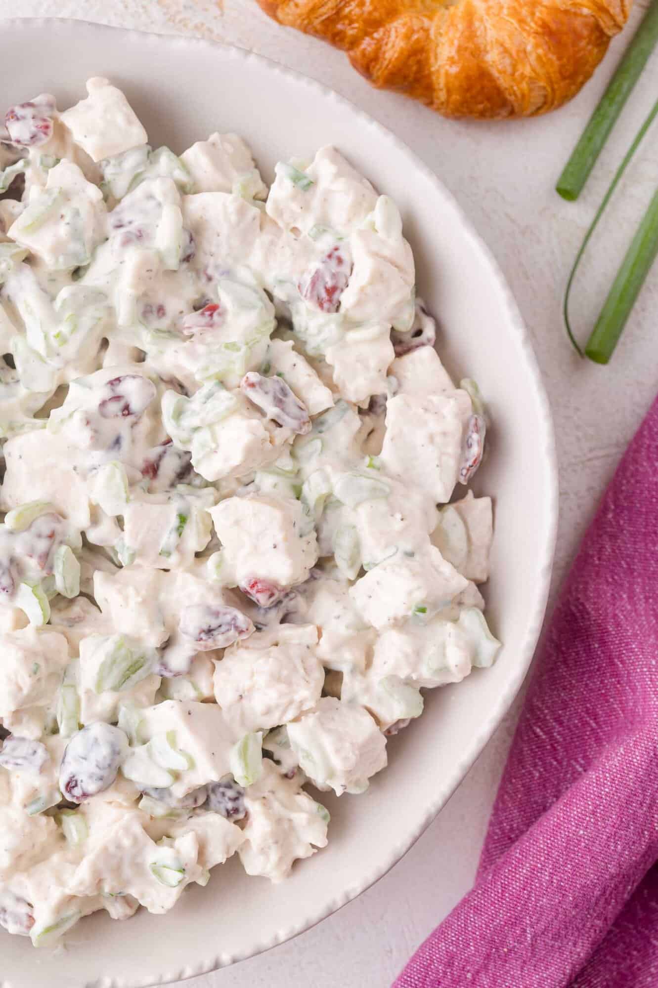 A bowl of creamy cranberry chicken salad with diced chicken, celery, and cranberries, placed on a white surface next to a croissant and green onions.