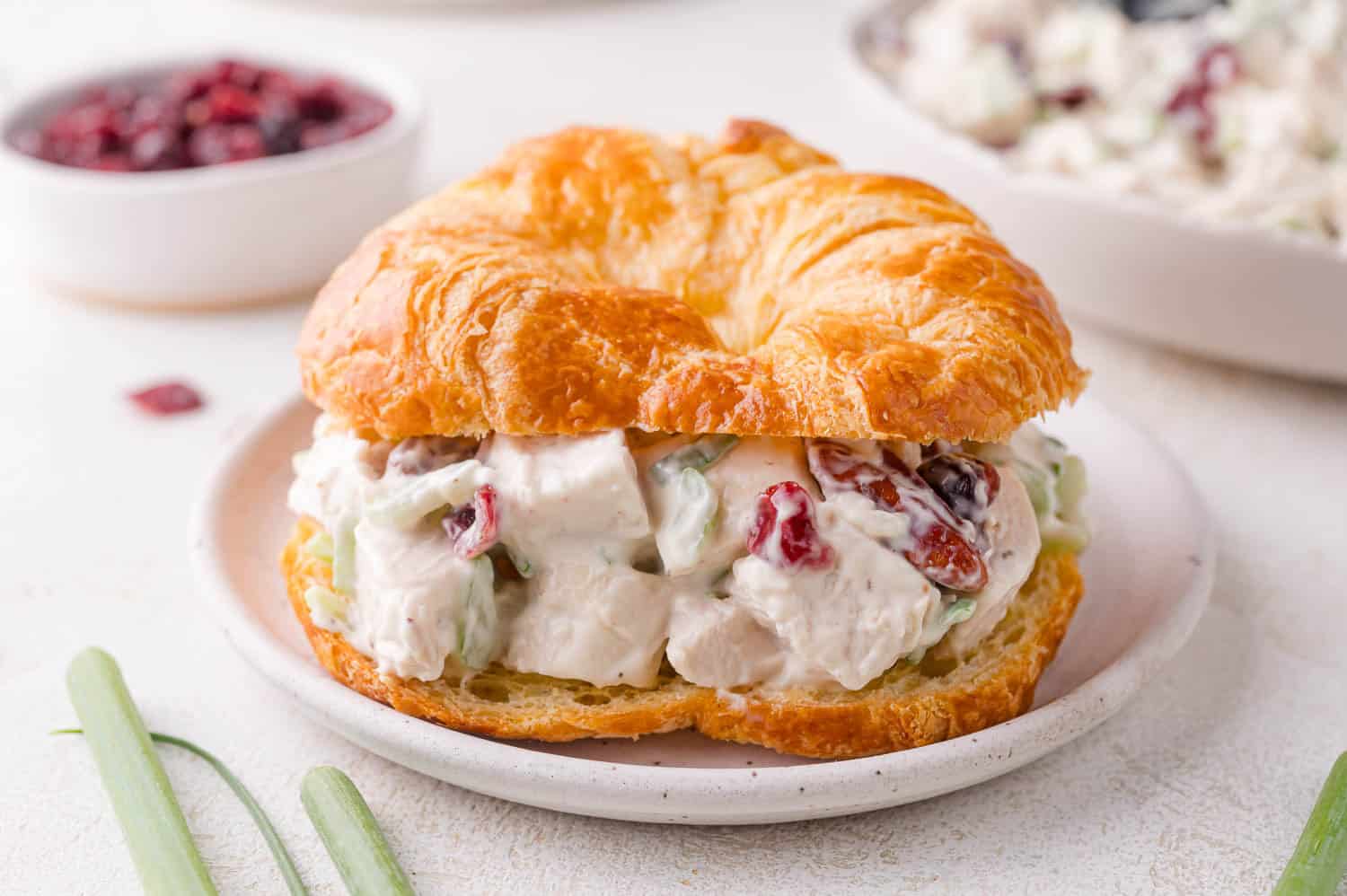 A croissant sandwich filled with chicken salad, diced celery, and dried cranberries on a white plate.