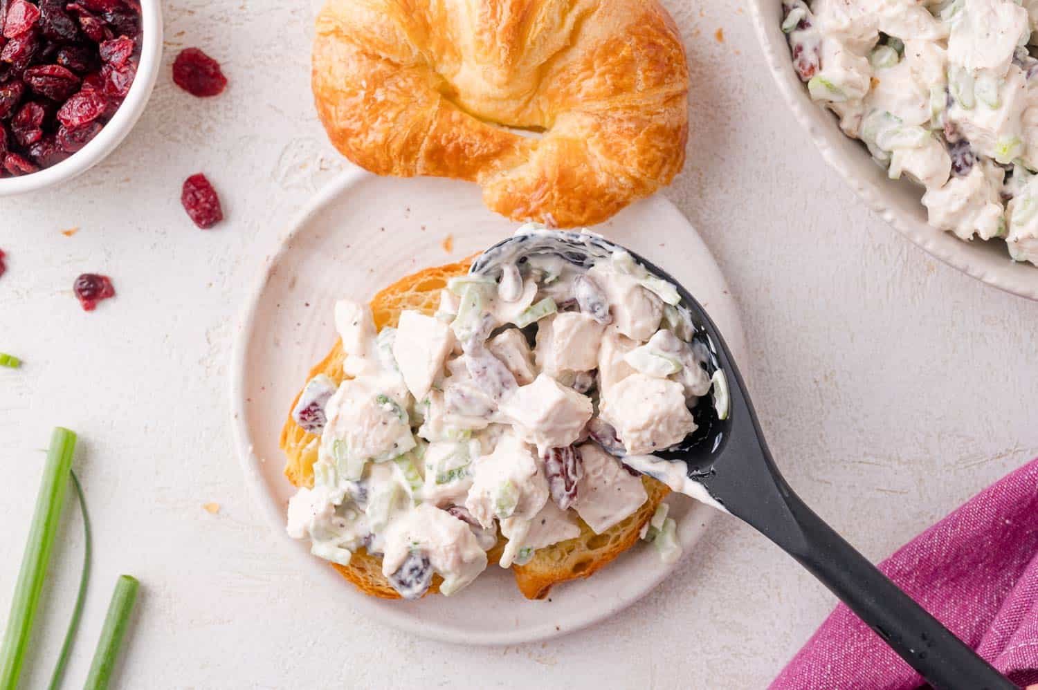 A croissant on a plate being topped with chicken salad containing diced chicken, celery, and dried cranberries; ingredients and a bowl are nearby.
