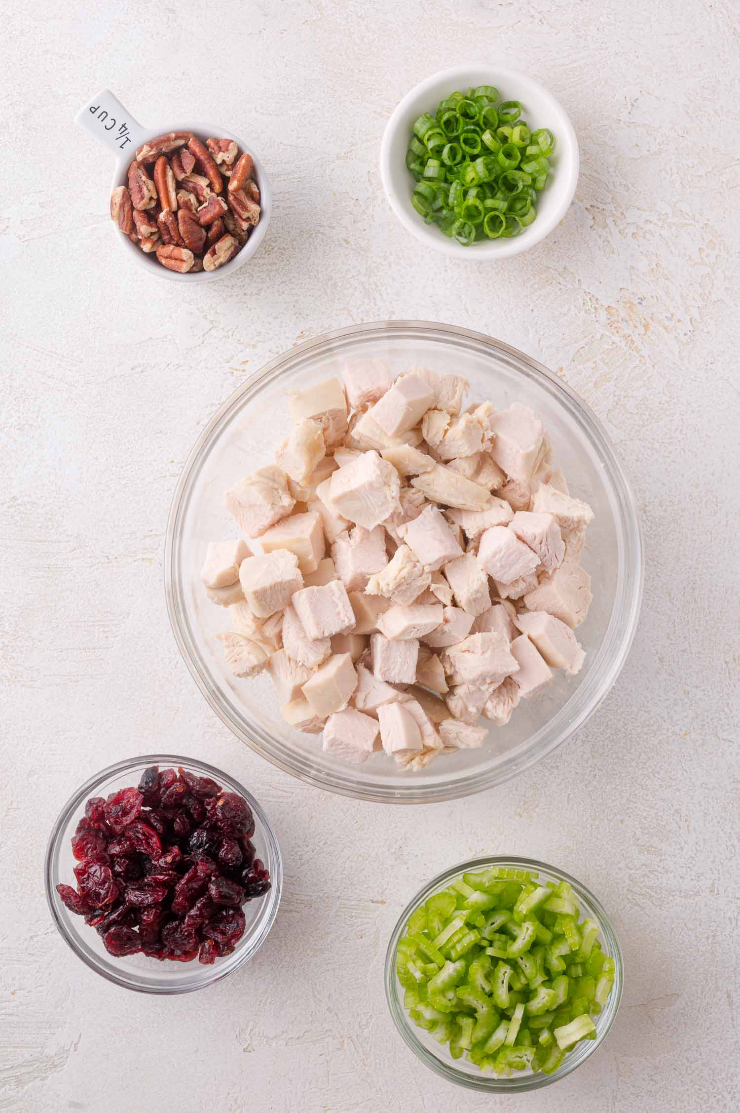 A glass bowl of cooked, cubed chicken surrounded by small bowls of chopped celery, green onions, dried cranberries, and pecans on a light surface.