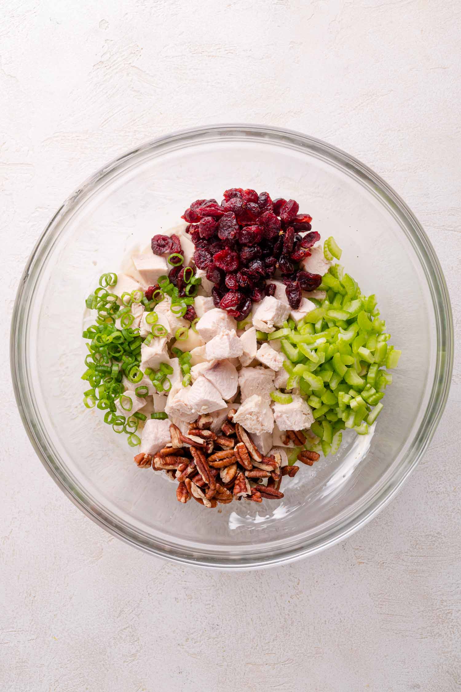 A glass bowl containing chopped cooked chicken, celery, green onion, dried cranberries, and pecans arranged in separate sections.