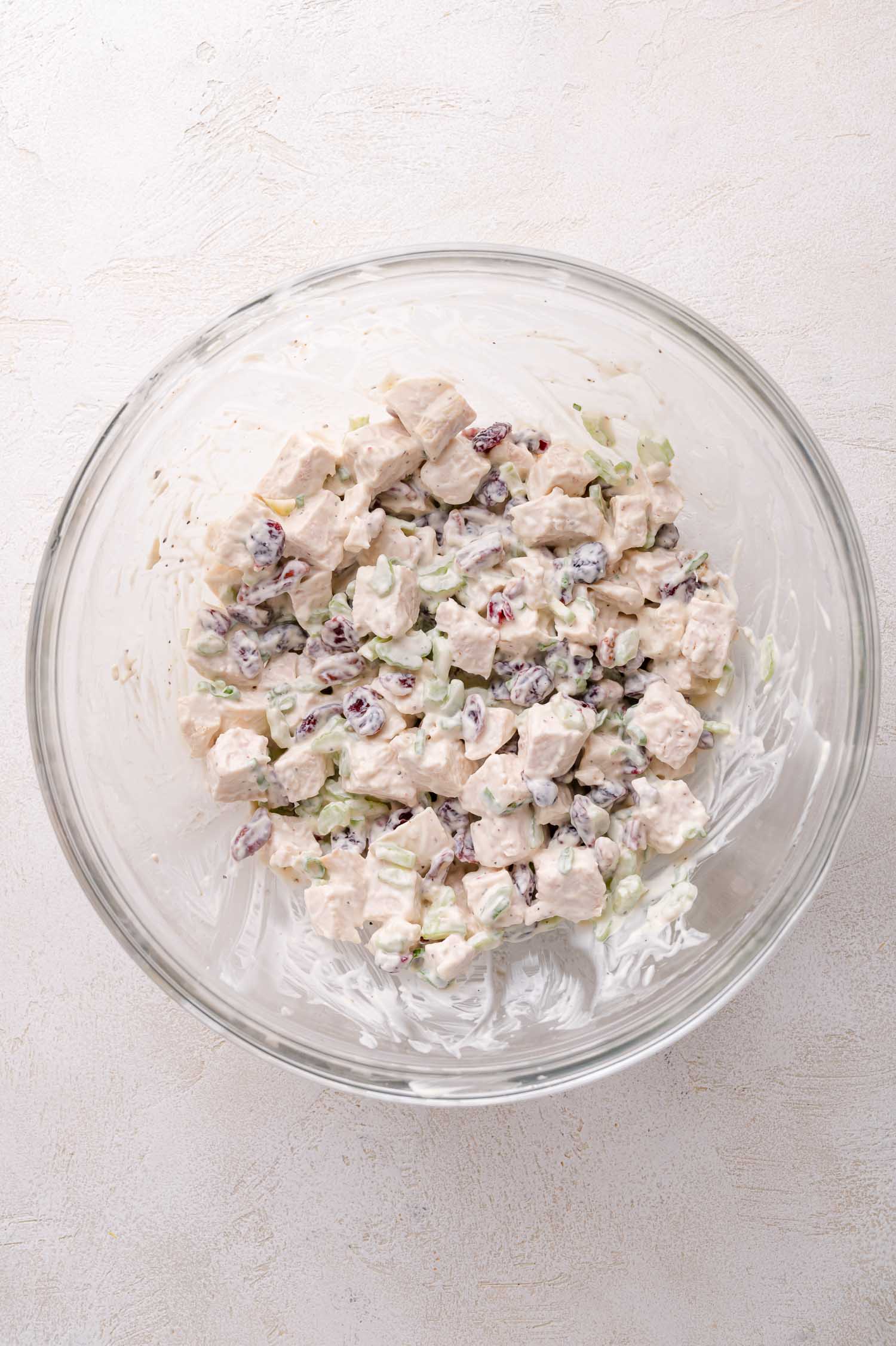 A glass bowl containing a mixture of diced chicken, celery, dried cranberries, and creamy dressing, set on a light-colored surface.
