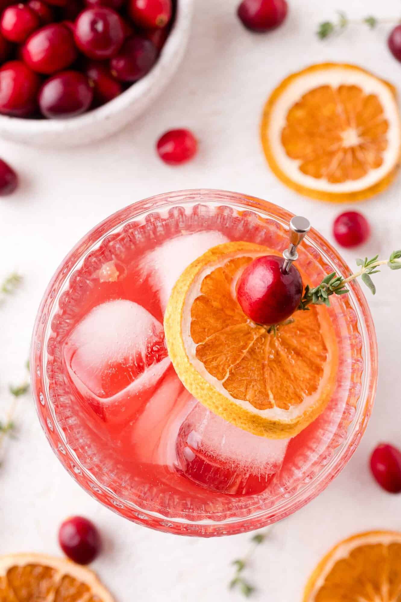 A glass of cranberry thyme spritzer with ice, garnished with a dried orange slice, a fresh cranberry, and a sprig of thyme. Sliced oranges and cranberries are scattered nearby.