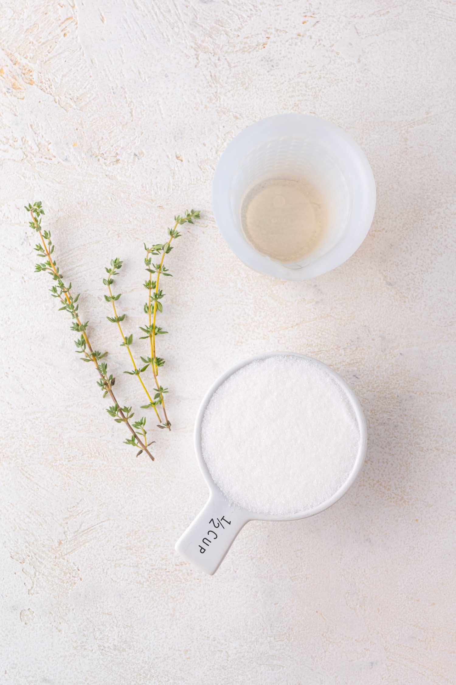 A half-cup of granulated sugar, a small clear cup of liquid, and two sprigs of fresh thyme arranged on a light surface.