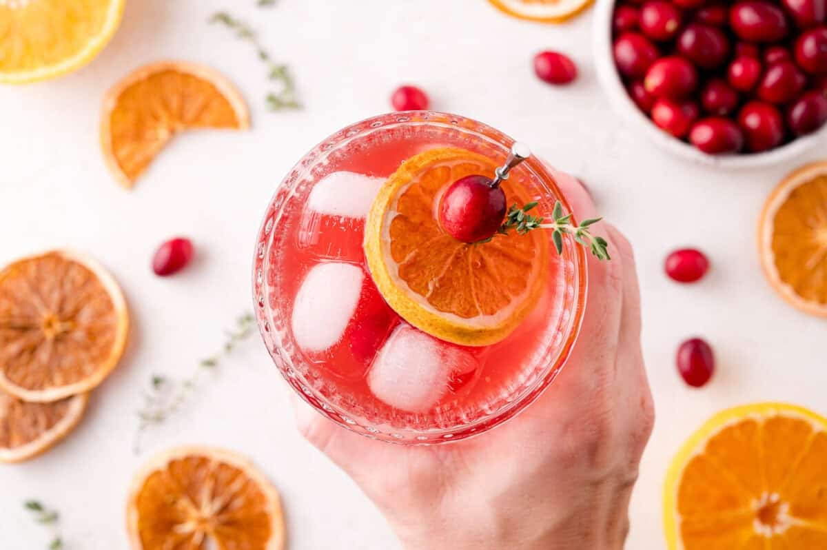 A hand holds a glass of cranberry thyme spritzer with ice, garnished with a dried orange slice, a cranberry, and a sprig of thyme. Dried oranges and cranberries are scattered on the table.