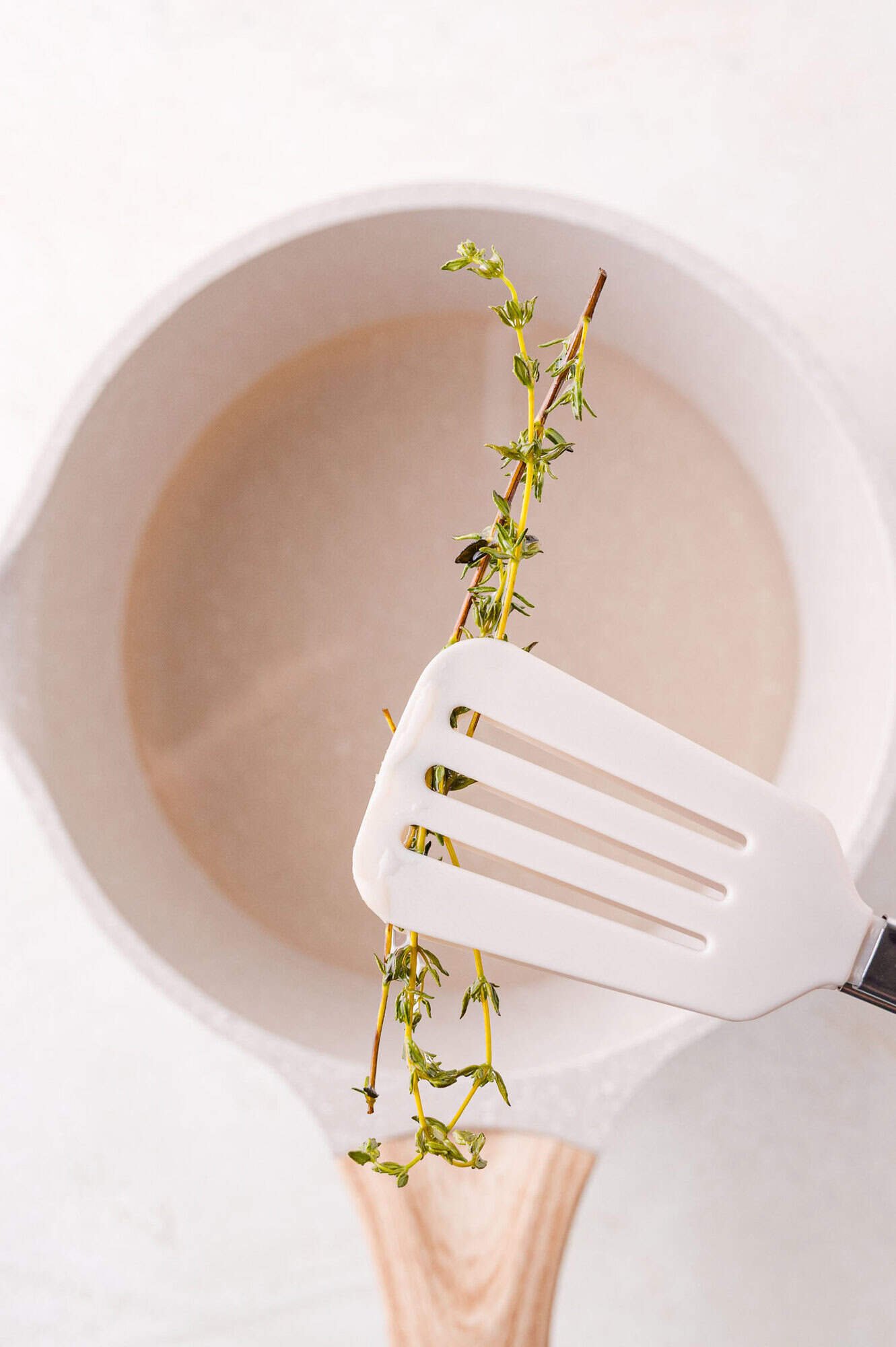A white spatula holds a sprig of fresh thyme over a white saucepan filled with simple syrup.