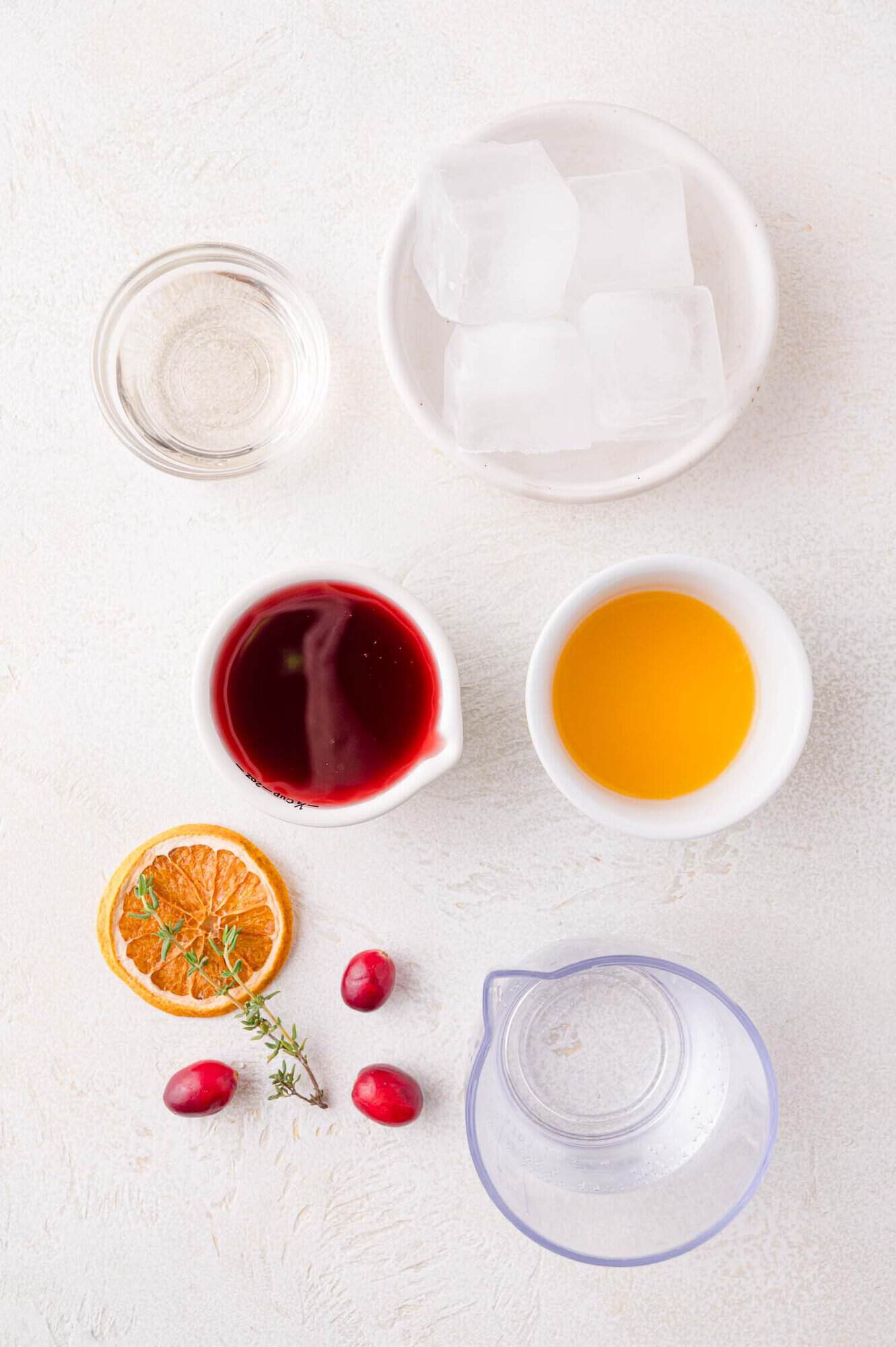 Top view of ingredients on a white surface: ice cubes, clear liquid, red liquid, orange liquid, a slice of dried orange, cranberries, thyme, and a clear liquid in a small pitcher.