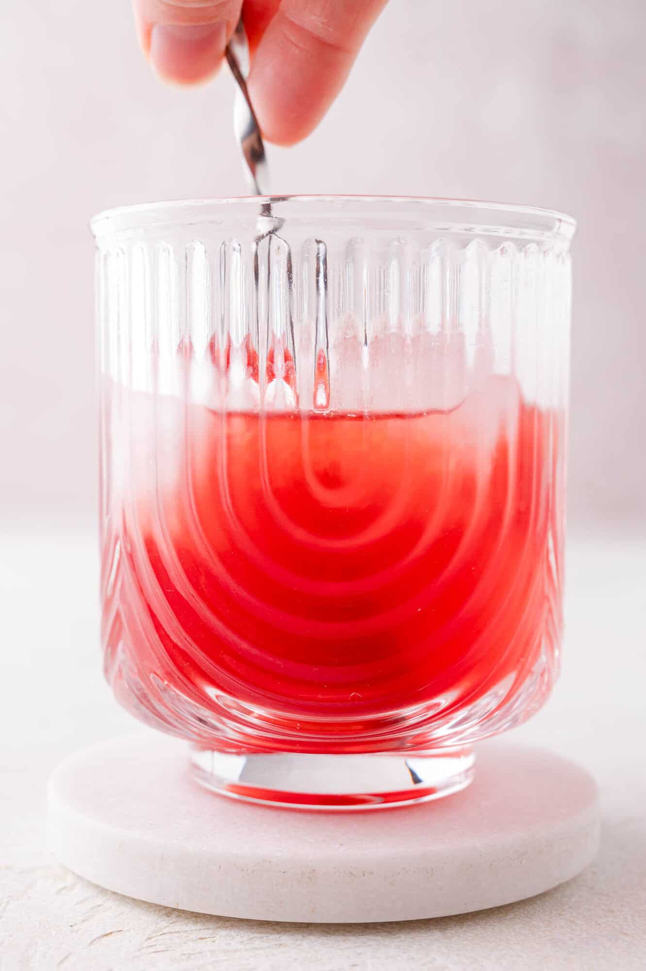 A hand stirs a red liquid in a clear glass with a textured pattern, placed on a round white coaster.