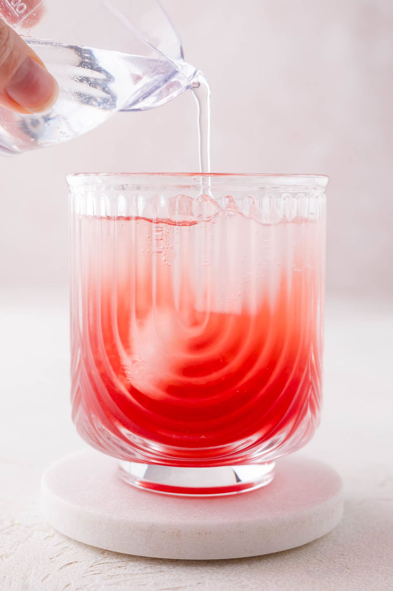 Clear liquid is being poured from a small glass into a textured glass containing a red beverage, causing the colors to mix.