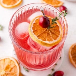 A glass of cranberry thyme spritzer garnished with a dried orange slice, cranberry, and sprig of thyme on a pick, with ice cubes and dried orange slices nearby.