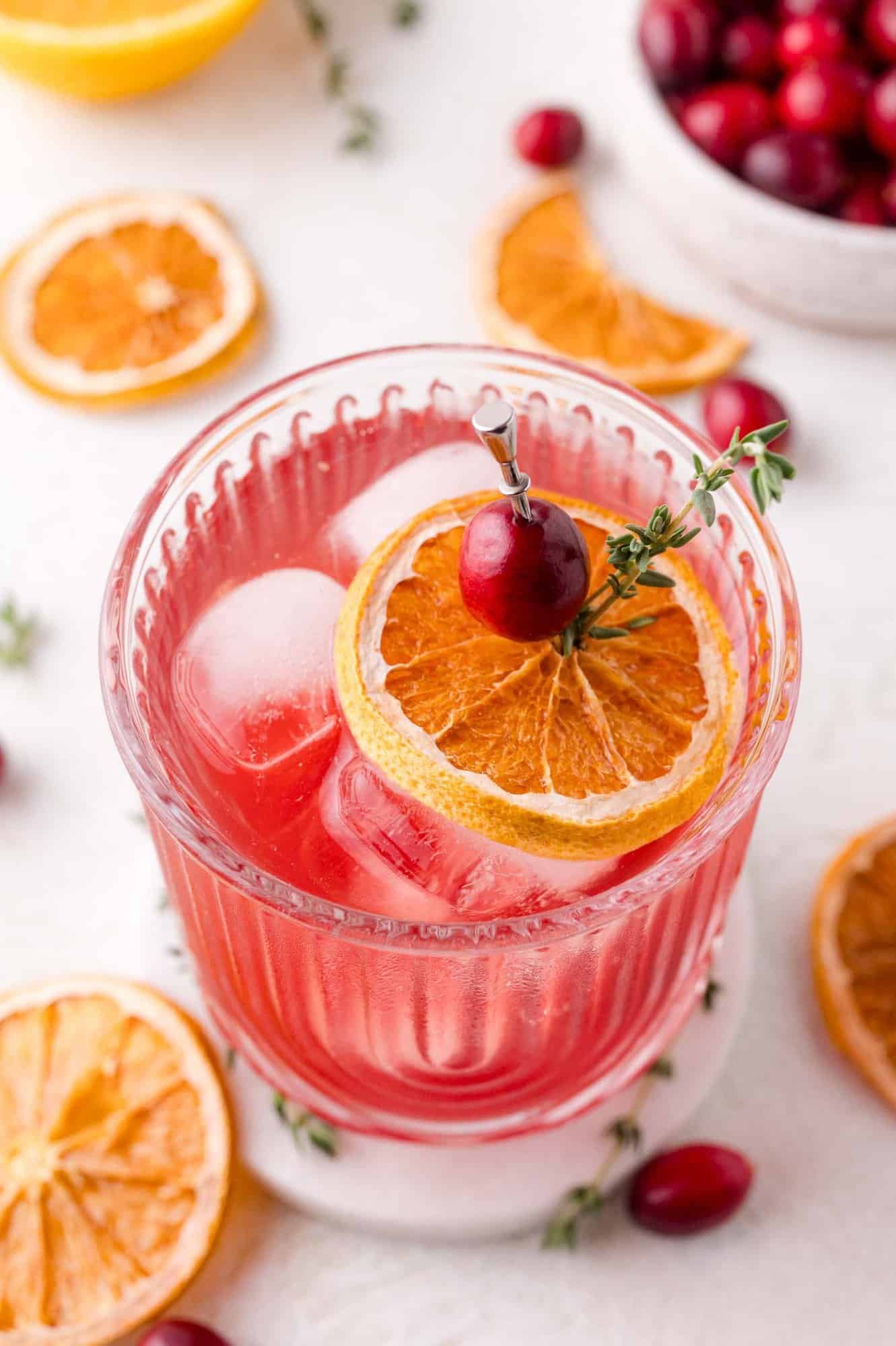 A glass of cranberry thyme sprtizer with ice, garnished with a dried orange slice, a cranberry, and a sprig of thyme, sits on a white coaster. Dried oranges and cranberries are scattered around.