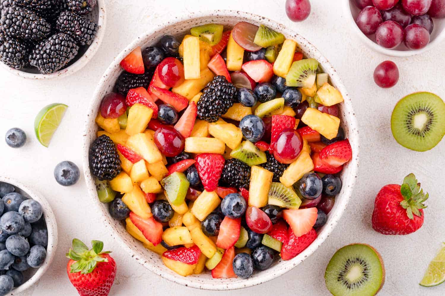 A bowl of mixed fruit salad with pineapple, strawberries, blueberries, blackberries, grapes, and kiwi, surrounded by small bowls of berries and slices of fruit on a white surface.