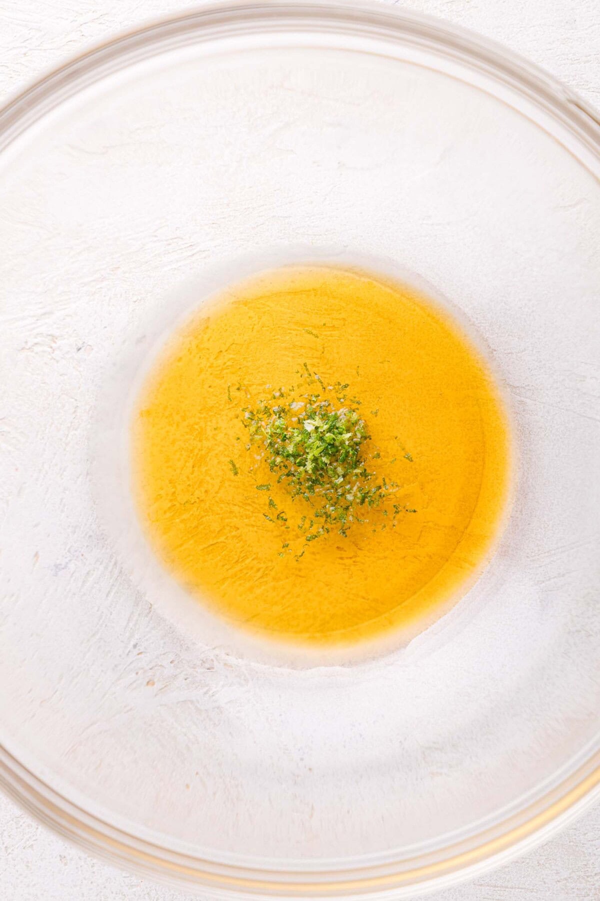 A clear glass bowl containing a mixture of yellow liquid and finely chopped green herbs on a white surface.