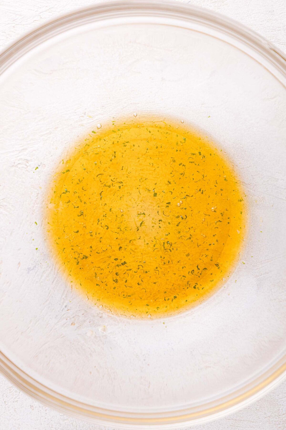 Clear glass bowl containing a small amount of yellow liquid with green specks of lime zest sitting on a white surface.