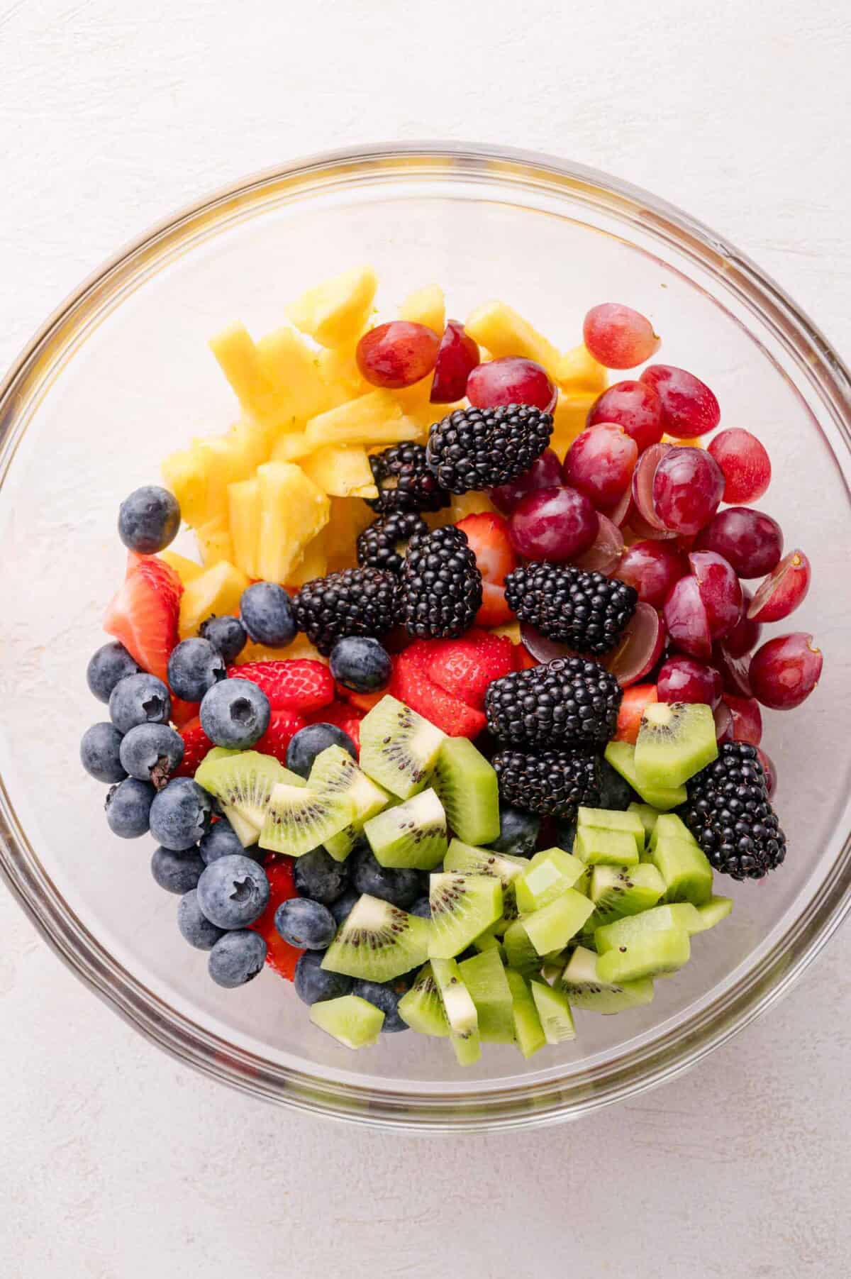 A glass bowl filled with sliced kiwi, blackberries, blueberries, strawberries, pineapple chunks, and red grapes on a white surface.