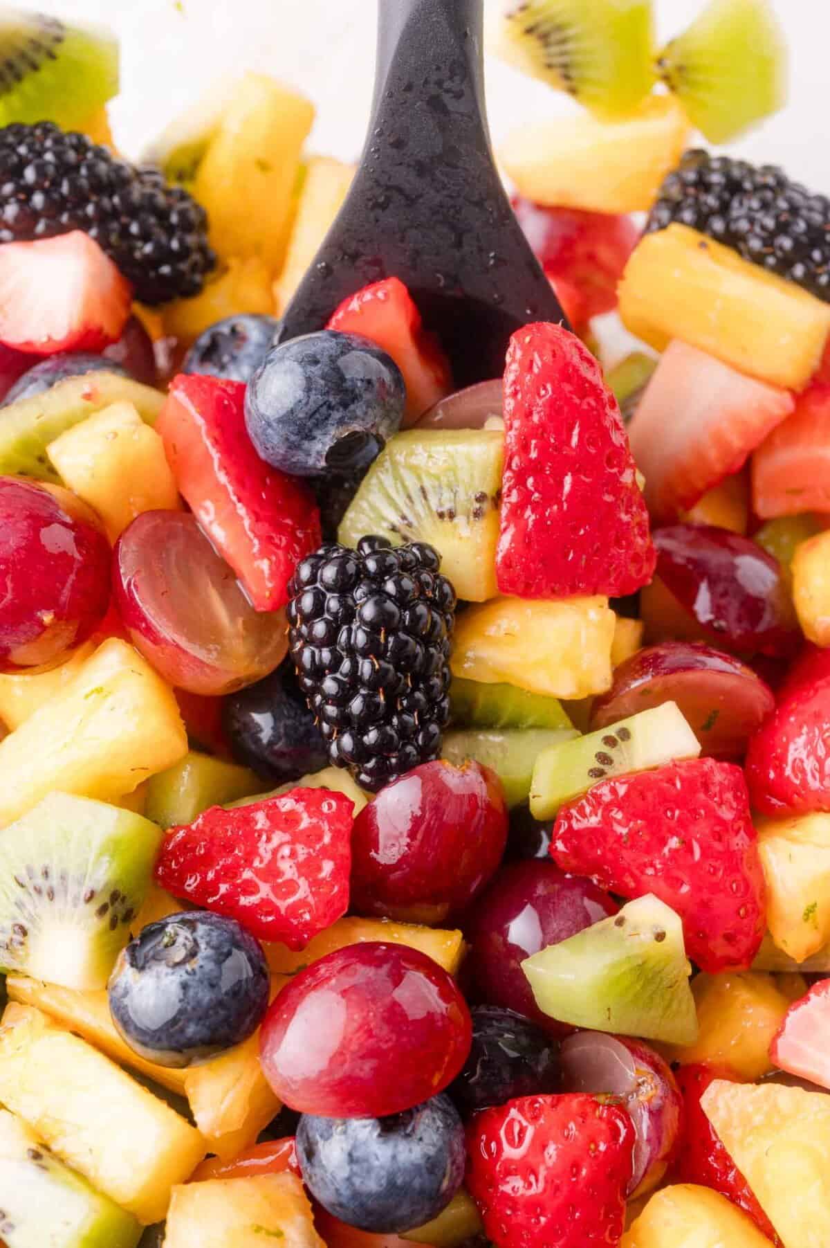 A close-up of a fruit salad with strawberries, kiwi, pineapple, grapes, blackberries, and blueberries, with a black spoon in the mix.