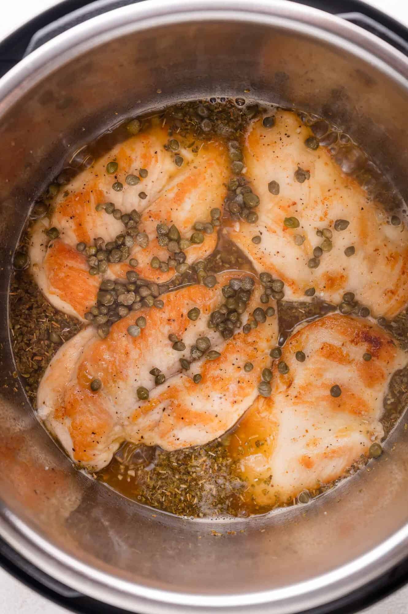 Four pieces of cooked chicken breast with capers and seasoning in a pot with broth.