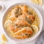 A bowl of cooked angel hair pasta topped with seared chicken piccata and a lemon-caper sauce, garnished with lemon slices.
