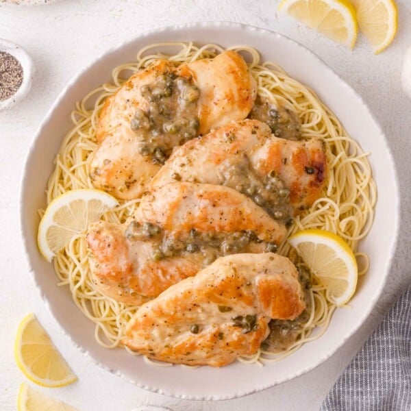 A bowl of cooked angel hair pasta topped with seared chicken piccata and a lemon-caper sauce, garnished with lemon slices.