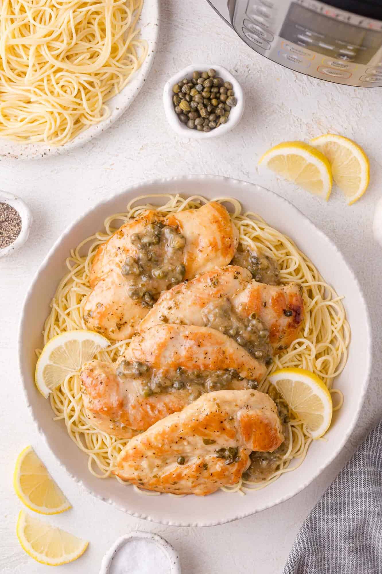 A bowl of cooked angel hair pasta topped with chicken breast in a caper sauce, garnished with lemon slices, with additional spaghetti, capers, lemon slices, and an Instant Pot nearby.
