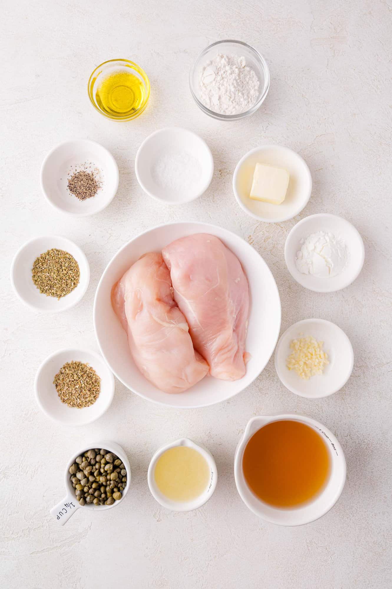 Raw chicken breasts in a bowl surrounded by small dishes of flour, oil, spices, butter, broth, lemon juice, minced garlic, and capers on a white surface.