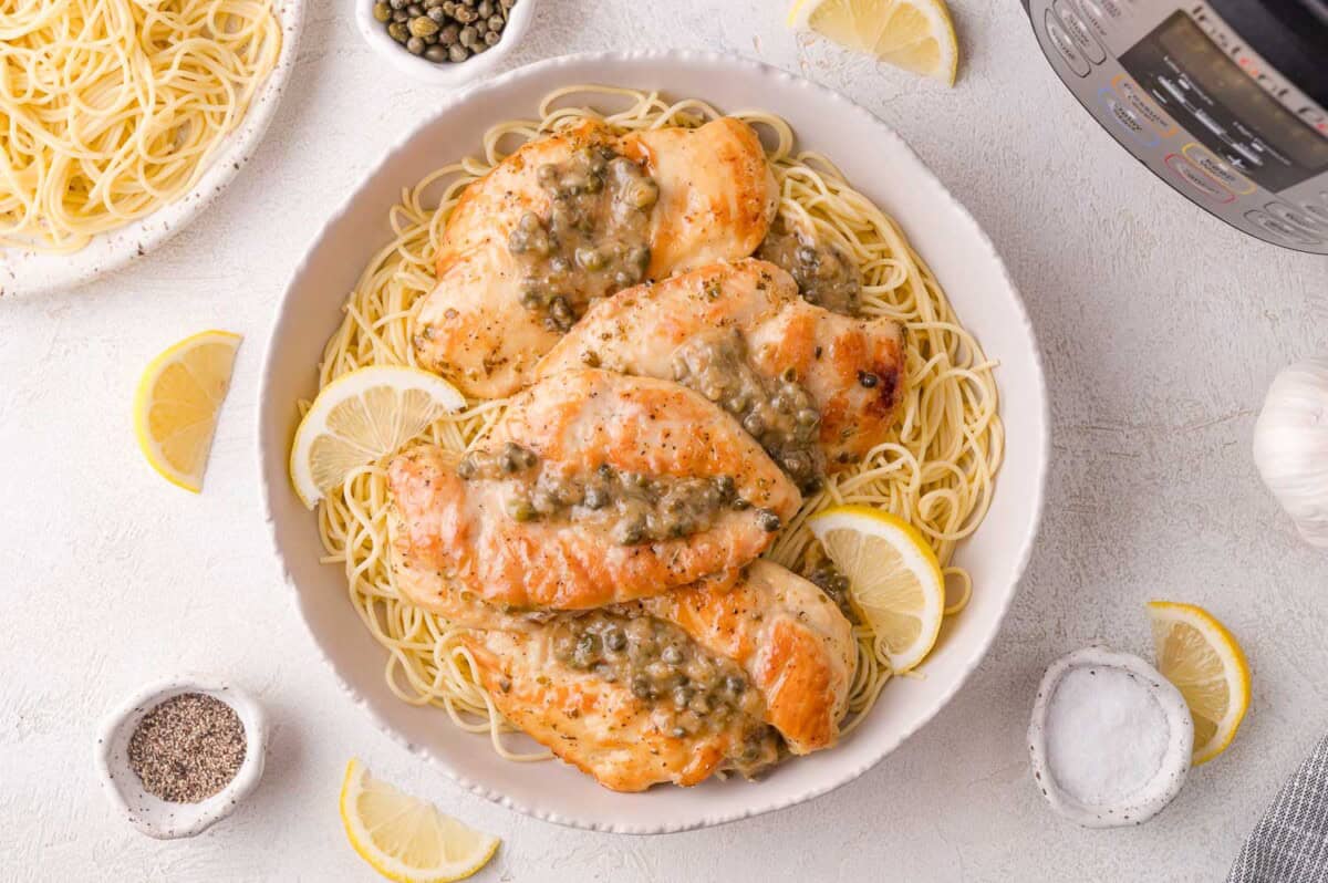 A bowl of angel hair pasta topped with cooked chicken breasts in a caper sauce, garnished with lemon slices, with seasonings and an Instant Pot on the side.