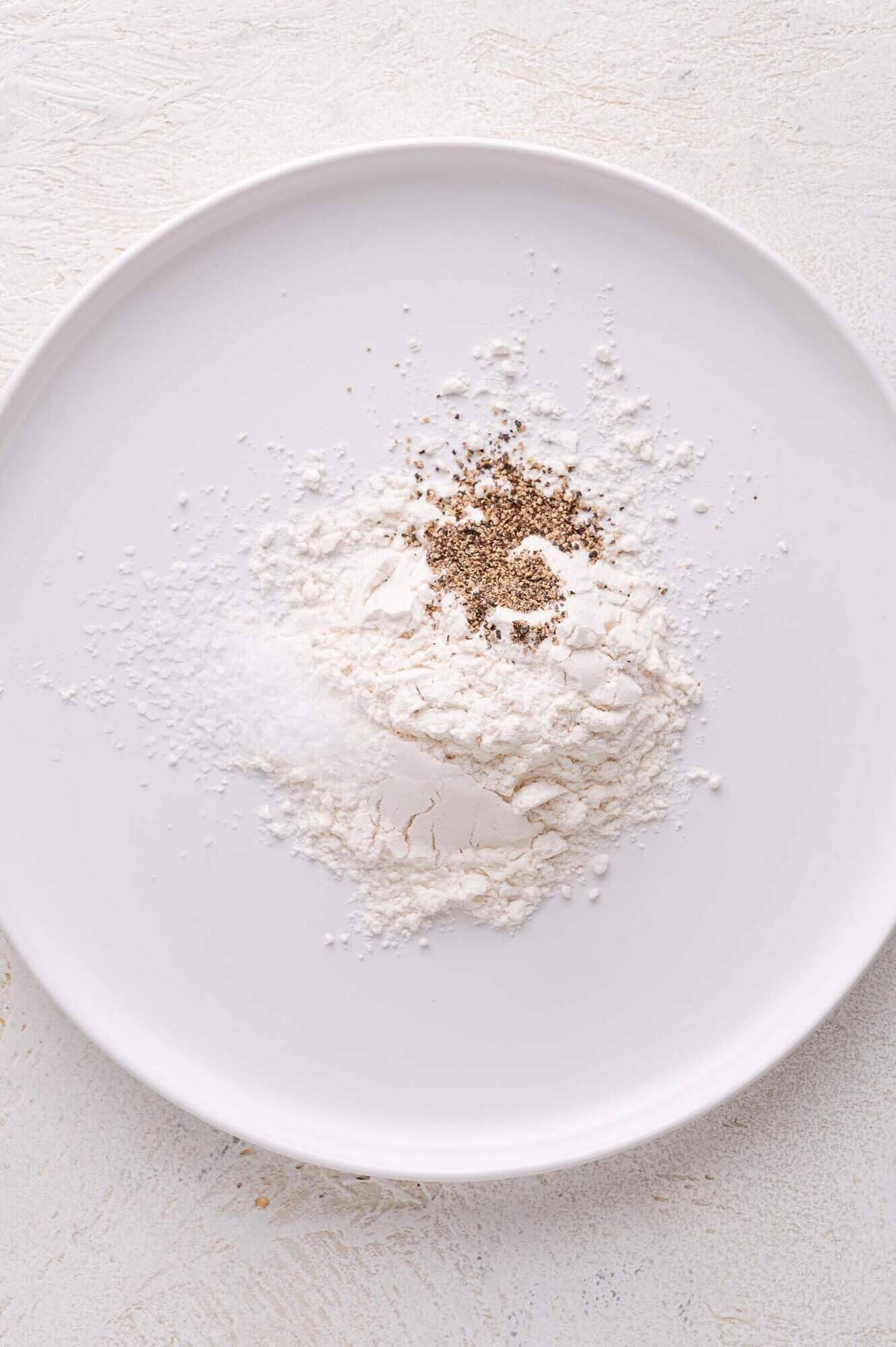 A white plate with small piles of flour, salt, and ground black pepper on a light-colored surface.
