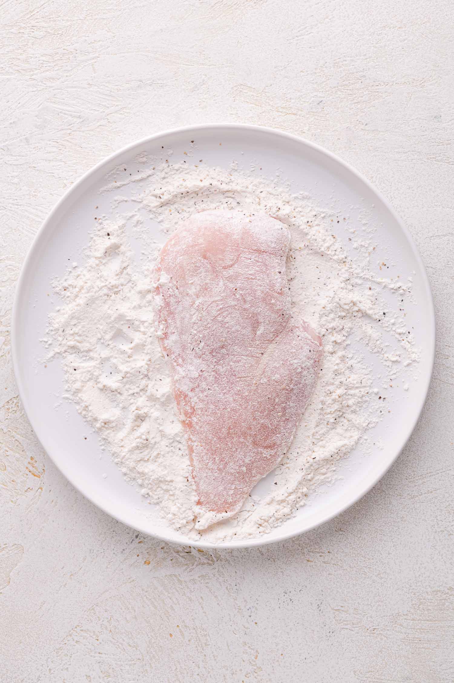 Raw chicken breast coated in flour and black pepper on a white plate, set on a light-colored surface.