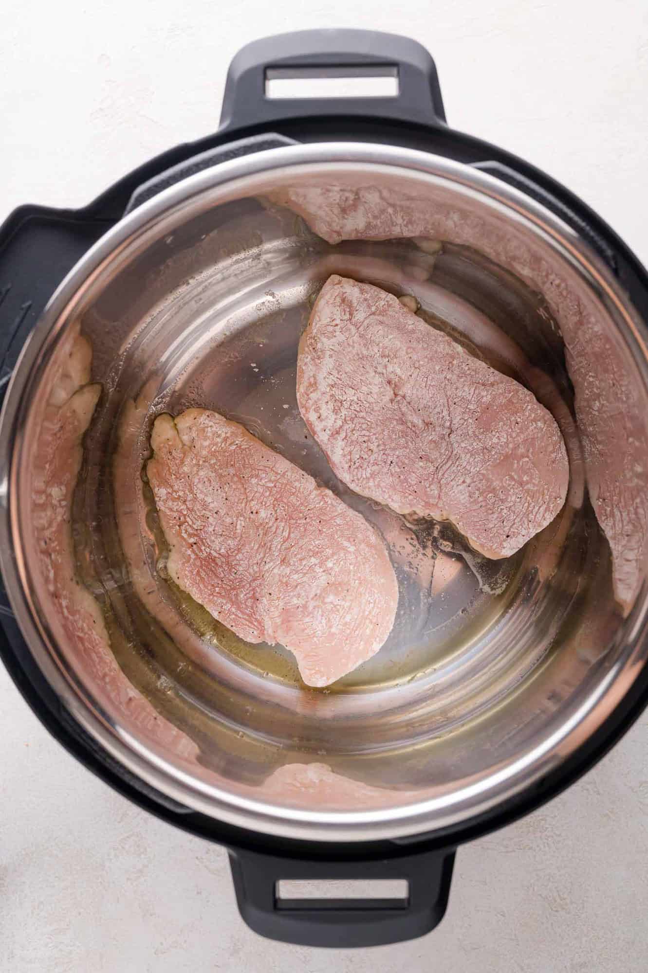 Two raw, floured chicken breasts placed inside the stainless steel inner pot of an electric pressure cooker.
