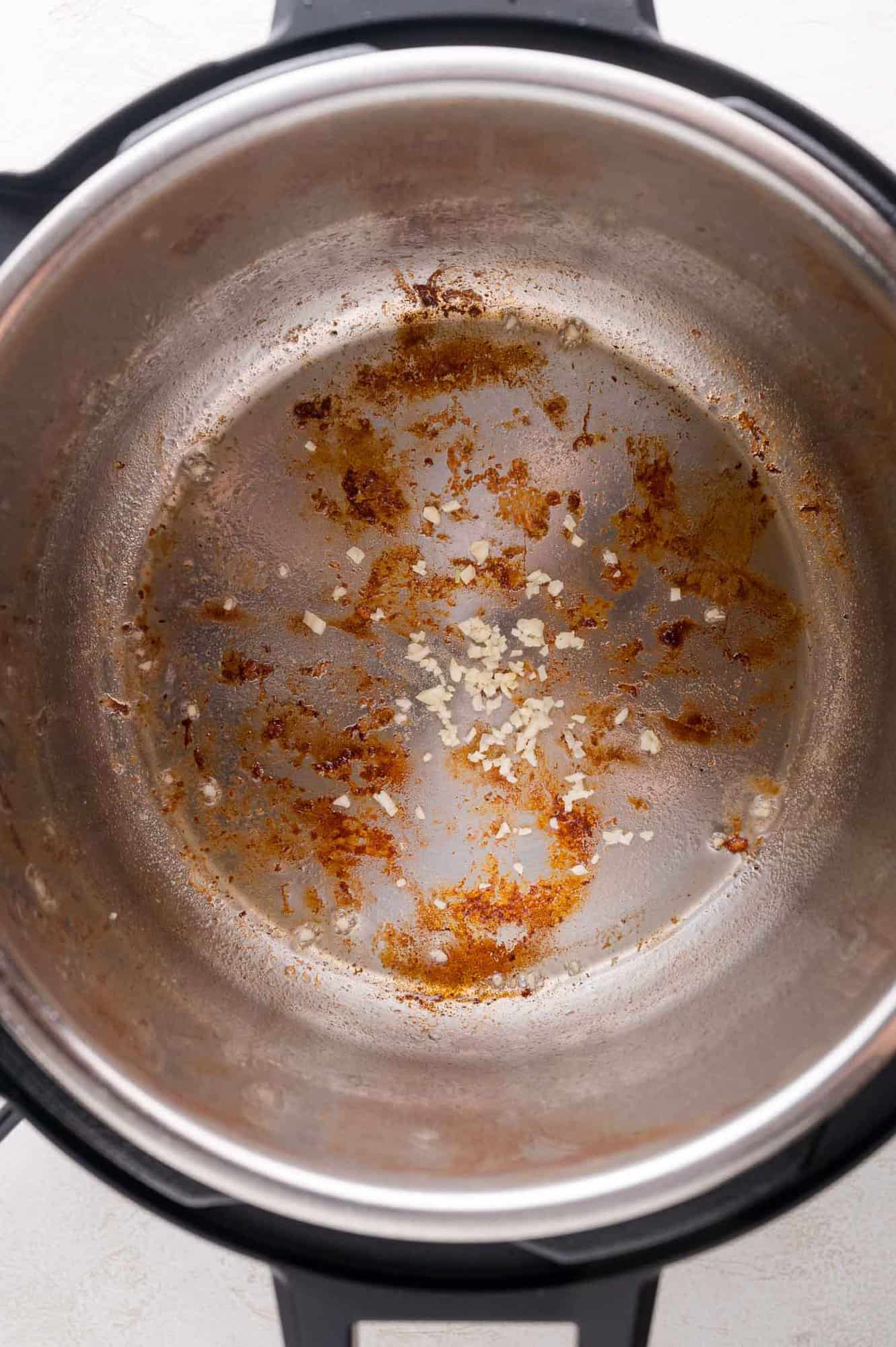 A close-up of the inside of a metal Instant Pot with browned bits and small pieces of sautéed garlic stuck to the bottom.
