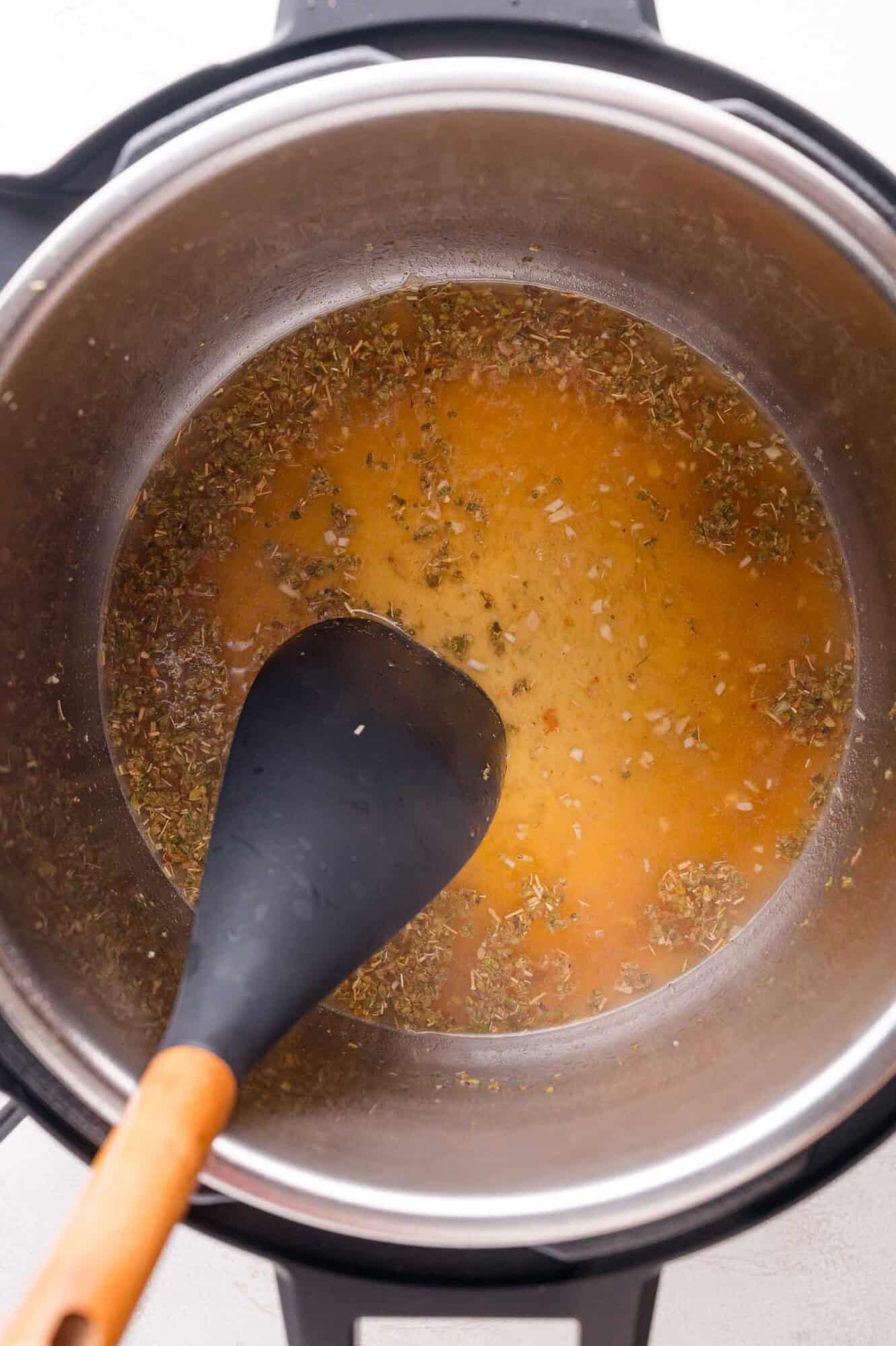 A black spatula stirs a seasoned broth in a stainless steel pot, with herbs and spices visible in the liquid.