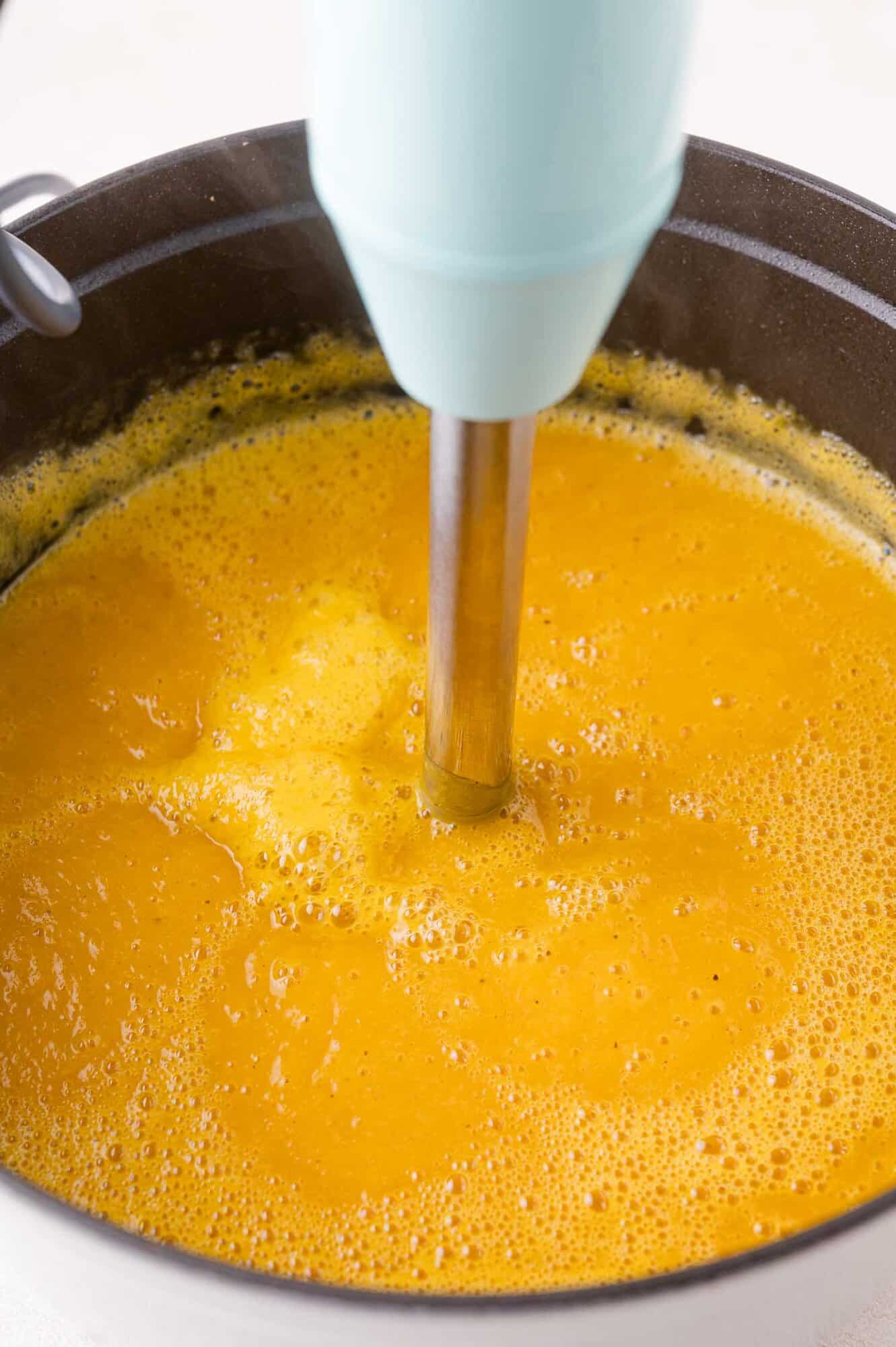 An immersion blender is blending kabocha squash soup in a black pot, creating a smooth and frothy texture.